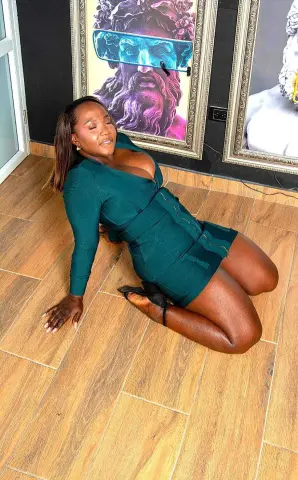 Smiling Black woman in a dark green long-sleeve mini dress sitting on a wood floor in a modern room.