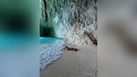 EvelynMills lies naked on a white sand beach next to the clear blue water of a sea cave.