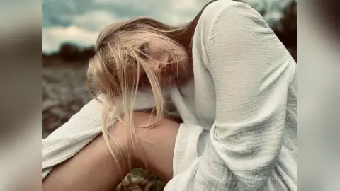 Blonde woman in a white linen outfit sitting in a dreamy outdoor field under a cloudy sky.