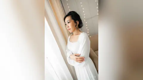 Elegant woman in a white long-sleeve dress looking out a window in a bright, dreamy indoor setting.
