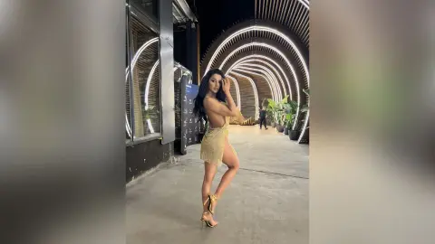 NicoleBella poses outdoors in a gold sequin mini dress with long dark hair, standing on a concrete walkway near a curved lit tunnel.