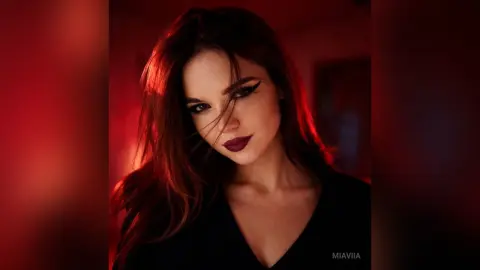 Moody portrait of a woman with red hair and dark makeup wearing a black outfit in dramatic red lighting.