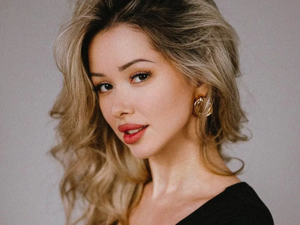 Blonde woman with gold hoop earrings wearing a black top in a soft studio portrait setting.