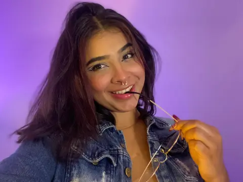 Smiling young woman in a denim jacket holding glasses against a purple studio background.
