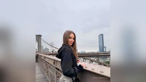 EvelynMills poses on a bridge walkway wearing a black hooded sweatshirt and patterned shorts against a city skyline.