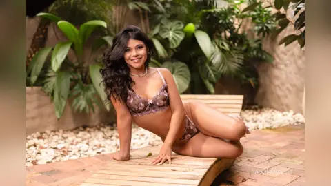 Smiling woman in floral lace lingerie relaxing on a wooden lounge chair in a lush tropical garden setting.