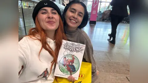 Two smiling women in casual outfits holding an Amazing Spider-Man comic book in a bright indoor setting.