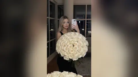 Blonde woman in a black dress holding a massive bouquet of white roses in a modern luxury apartment.