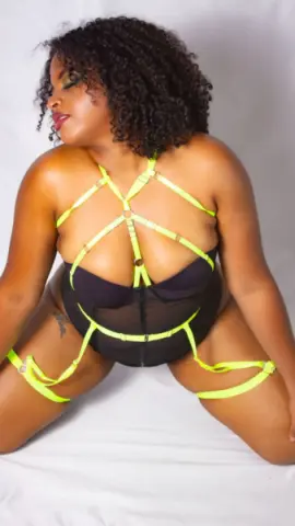 Curvy woman with curly hair in black lingerie and neon yellow harness bodysuit posing against a white background.