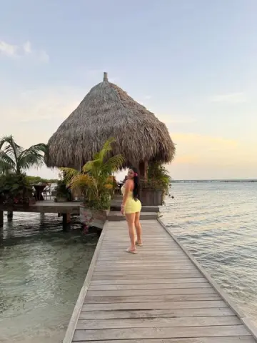 Woman in a yellow sundress walking on a wooden pier toward a tropical thatched hut at sunset.