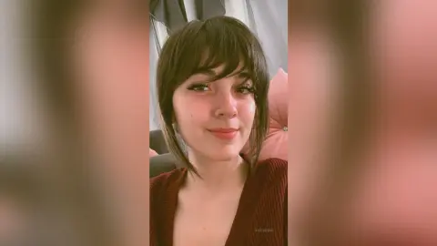 Smiling young woman with dark bangs wearing a cozy red sweater in a soft indoor setting.