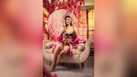 Glamorous woman in pink silk robe and black lingerie sitting on a vintage sofa amidst cherry blossom decor.