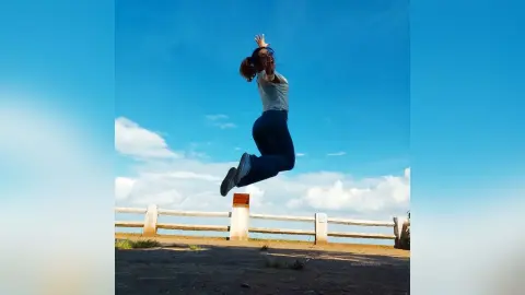 Energetic young woman in a white t-shirt and blue jeans jumping joyfully against a bright blue sky.