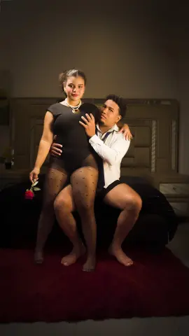 Romantic couple in intimate pose wearing elegant evening attire on a bed in a dimly lit room.
