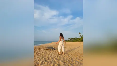 Young woman in white linen trousers walking on a sunny tropical beach towards the ocean waves.