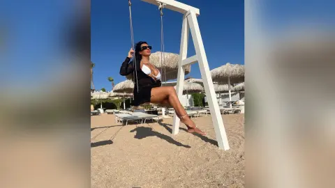 MissyJolie sits on a white swing wearing a black bikini and sunglasses against a clear blue sky.