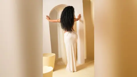 Elegant woman in a white maxi dress standing in a minimalist beige arched doorway with a serene mood.