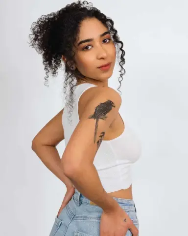 Young woman with curly hair in a white tank top and denim jeans posing against a white background.