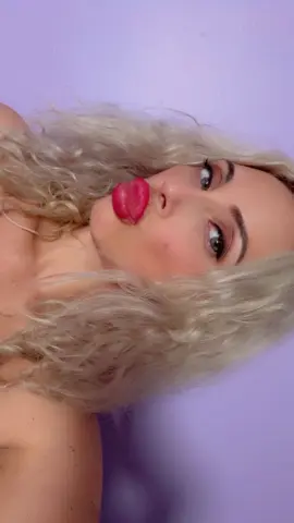 Close-up portrait of a blonde woman with wavy hair and red lipstick posing against a lavender background.