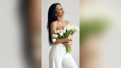 Serene woman in white trousers holding a large bouquet of white tulips against a minimalist studio background.