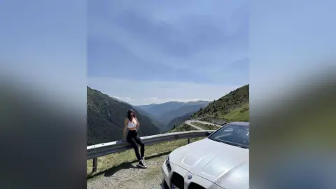 DyandraEve sits on a metal guardrail overlooking a deep mountain valley next to a white car.