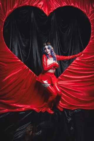 KimLennoxc poses in a red Harley Quinn cosplay outfit with long red gloves and a short skirt, framed by a large red heart shape.