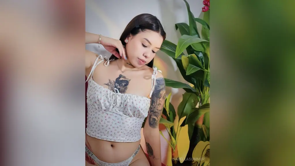 Young woman with tattoos in a white floral sundress posing near green houseplants in soft sunlight.