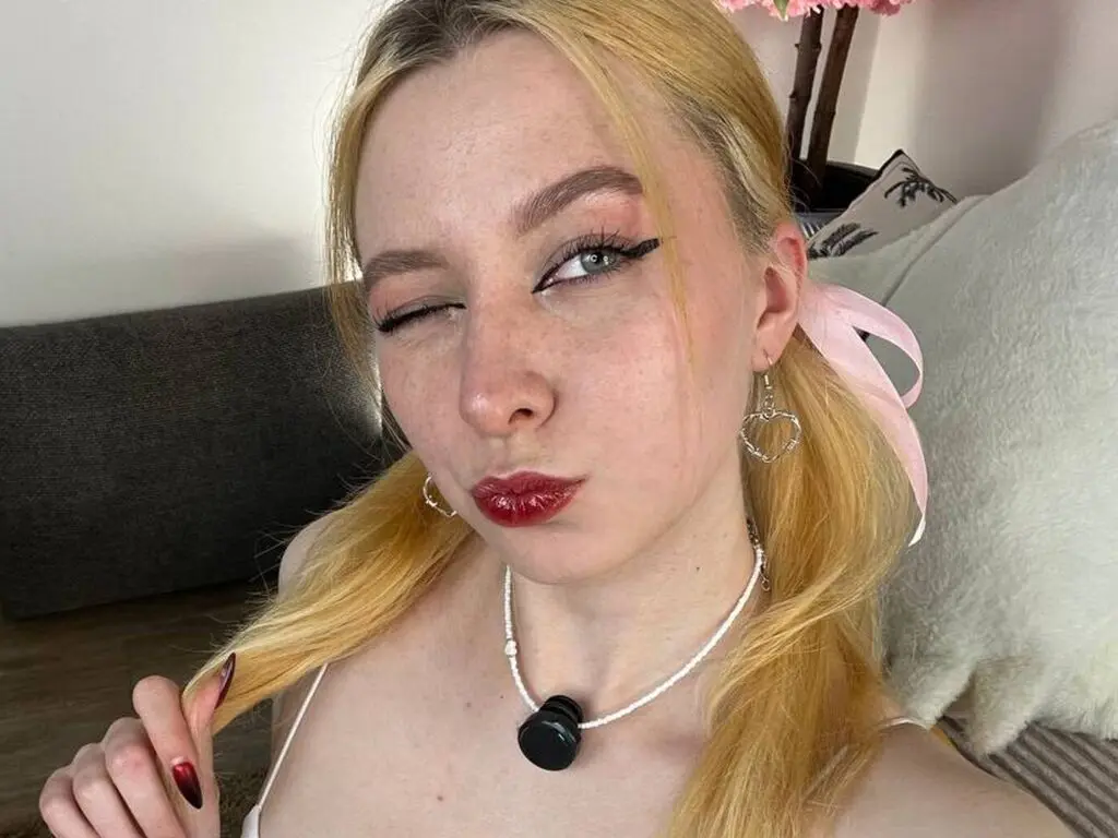 Blonde woman with red lipstick and white necklace winking in a cozy indoor setting.