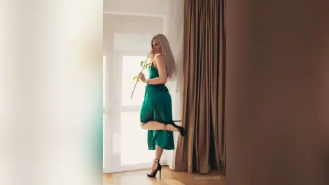 Elegant blonde woman in a green satin dress holding a rose by a bright window in a dreamy indoor setting.
