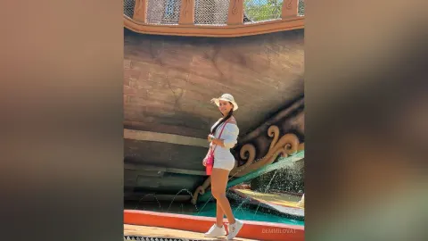 DemmiLovat — DemmiLovat stands in a white outfit and sun hat in front of a large wooden ship structure.