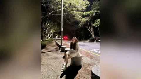 Young woman in a white sweater sitting on a concrete bollard on a tree-lined street at night.
