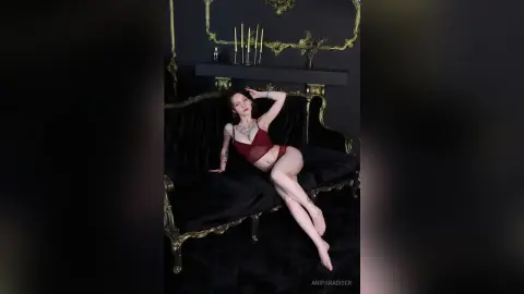 AniParadiser reclines on a black velvet sofa wearing a red lace bodysuit, showing off her tattoos and long legs against a dark, gold-trimmed background.