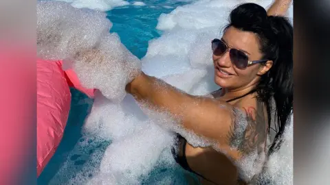MissyJolie smiles while relaxing in a bubbly white foam bath, showing her tanned skin and shoulder tattoo.