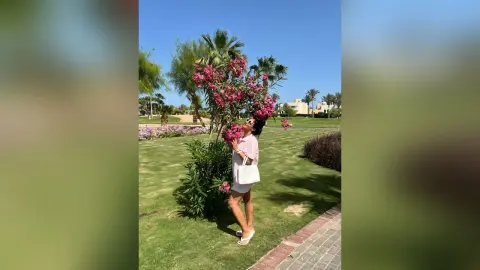 MissyJolie stands on a green lawn next to a flowering pink bush, wearing a white sleeveless top and white shorts.