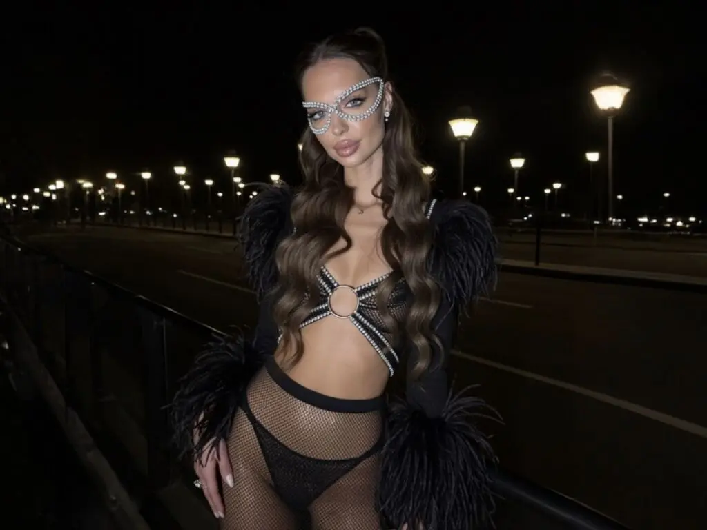 Glamorous woman in a black feathered cutout bodysuit posing at night by a city street with glowing lamp posts.