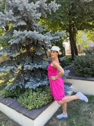 Woman in a pink summer dress and white sun hat posing by a blue spruce tree in a sunny garden.