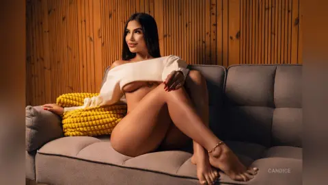 Glamorous woman in a white off-the-shoulder top relaxing on a grey sofa against a wooden slatted wall.