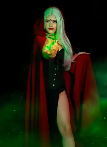 AlicceGray poses in a black corset and red hooded cape, holding a glowing green light in her hand against a dark, misty background.