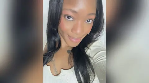 Smiling Black woman with long dark hair wearing a white ribbed top in a bright indoor setting.