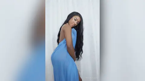 Beautiful woman in a flowing blue backless dress posing against a white curtain in soft lighting.