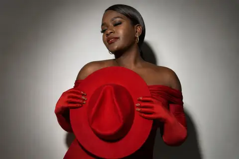 Elegant Black woman in a red off-the-shoulder dress and gloves holding a matching wide-brimmed hat against a gray wall.