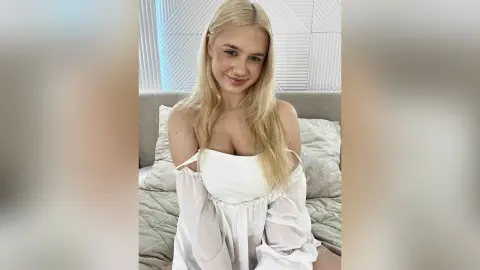 Smiling blonde woman in a white off-the-shoulder blouse sitting on a bed in a bright, airy bedroom.