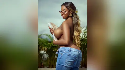 Curvy woman in denim jeans and sunglasses holding a book outdoors in a sunny tropical setting.