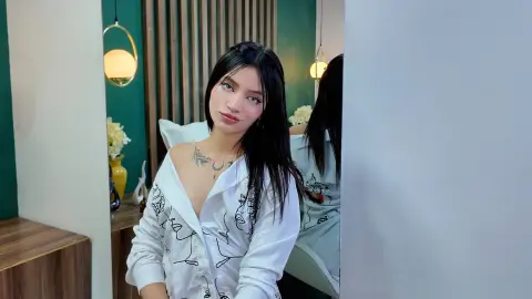 ScarlettPrescott poses in a white button-down shirt with black line art, showing her long black hair and chest tattoo against a green wall background.