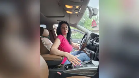 OpheliaDumas sits in the driver's seat of a car wearing a pink t-shirt and blue jeans, looking toward the camera with long black hair.