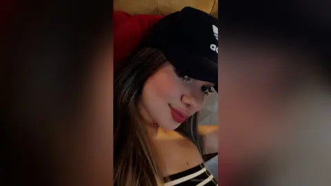 Smiling young woman in a black Adidas cap and white top posing for a cozy indoor selfie.