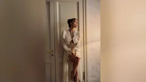 Elegant woman in a white lace sheer gown leaning against a door in soft, moody indoor lighting.