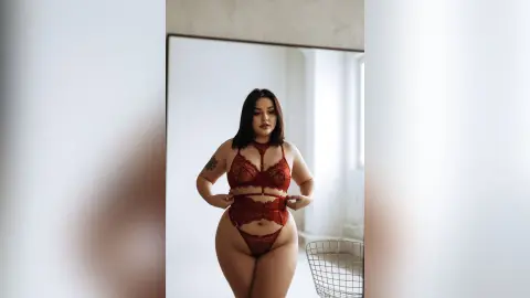 Curvy woman in red lace lingerie posing confidently in a bright, minimalist modern bedroom.