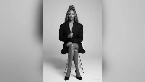 Elegant Black woman in a chic black blazer sitting on a chair against a minimalist studio background.