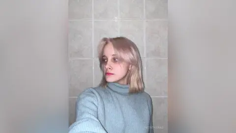 Blonde woman in a blue ribbed turtleneck sweater posing against a neutral tiled wall with a soft, moody gaze.